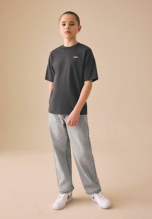 Dreng iført en sort T-shirt, lysevaske jeans og hvide sneakers, stående mod en ensfarvet beige baggrund.