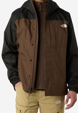 Homme portant une veste North Face marron et noire par-dessus une couche zippée marron et un pantalon North Face beige, mains partiellement visibles.