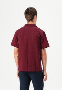 Chemise bourgogne à manches courtes avec motif en losanges texturé, col rabattable et ourlet droit. Le matériau semble léger et respirant.