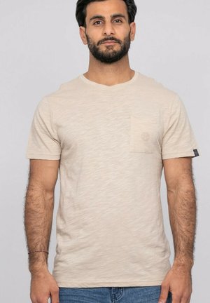 Homme barbu portant un T-shirt beige à manches courtes avec une poche poitrine, debout devant un fond gris clair uni.