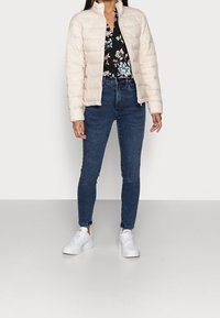 Světle béžová péřová bunda s vysokým límcem, v kombinaci s tmavě modrými skinny džínami a bílými teniskami, stojící na neutrálním pozadí.