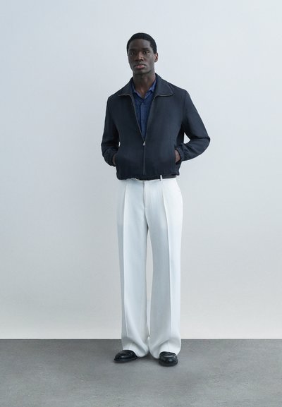 Homme debout, les mains dans les poches, portant une veste marine, une chemise bleue, un pantalon blanc à jambes larges et des chaussures noires, sur un fond uni.