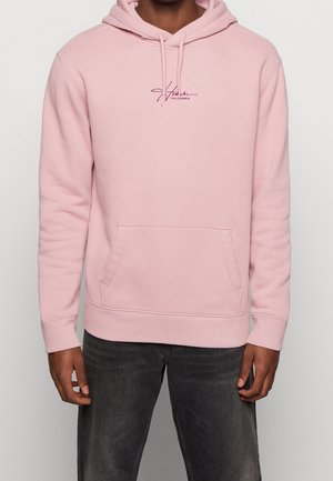 Man draagt een lichtroze hoodie met voorzak en klein "California" tekstlogo in donkerroze, gecombineerd met donkere spijkerbroek.