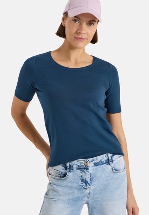 Femme portant un t-shirt bleu marine à manches courtes, un jean bleu clair et une casquette de baseball rose clair, debout avec une main sur la hanche.