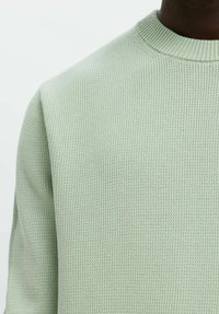 Zbliżenie na osobę w jasnolistnej, fakturowanej swetrze z okrągłym dekoltem na białym tle.