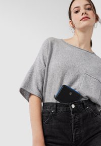 Maglione in misto lana grigio a maniche corte con tasca frontale, abbinato a jeans in denim neri a vita alta e a un piccolo portafoglio nero con accenti blu.