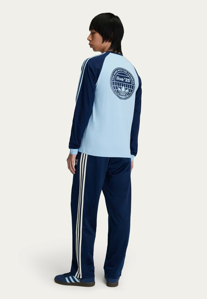 Maglia azzurra a maniche lunghe con maniche blu navy, dotata di una grafica sul retro; pantaloni blu navy con tre strisce bianche sui lati.