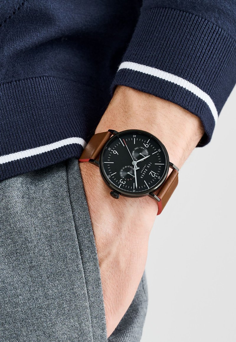 Reloj negro con tres subesferas, correa de cuero marrón, caja de acero inoxidable y marcadores de hora blancos, llevado en una muñeca contra ropa gris.