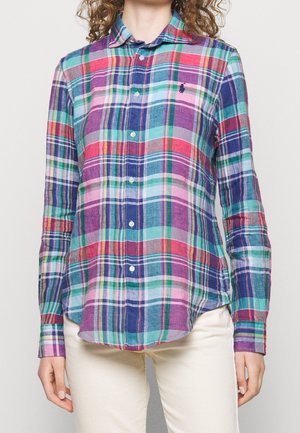 Femme portant une chemise à manches longues boutonnée avec des motifs à carreaux multicolores et un pantalon de couleur claire, debout devant un fond uni.