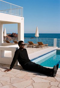 Homme portant des lunettes de soleil et des vêtements noirs assis au bord d'une piscine avec des chaises longues, des parasols et une vue sur l'océan en arrière-plan.