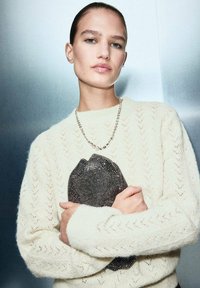 Pull en tricot blanc cassé avec un motif en chevron, doté de manches longues. Une pochette noire pailletée en forme de lèvres est tenue devant.