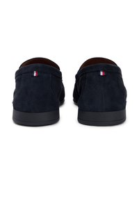 Σουέντ παπούτσια που φοριούνται χωρίς κορδόνια σε ναυτικό μπλε, με χαμηλή μαύρη καουτσούκ σόλα και μικρή τριχρωμία σημαία στο τακούνι. Λείο υφαντό, μινιμαλιστικός σχεδιασμός.