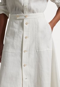 Vestido de lino blanco con cierre frontal de botones, dos bolsillos grandes y cintura ceñida con botones a juego. Tejido suave y texturizado.