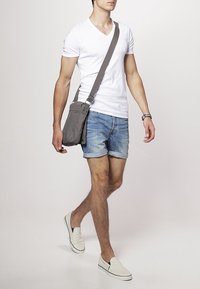 Bolso de hombro gris con correa ajustable, llevado por una persona con una camiseta blanca de cuello en V y pantalones cortos de mezclilla, caminando sobre un fondo neutro.