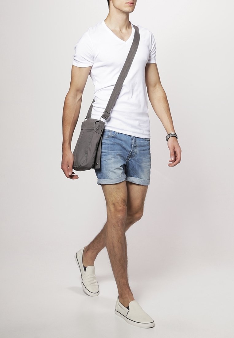 Bolso de hombro gris con correa ajustable, llevado por una persona con una camiseta blanca de cuello en V y pantalones cortos de mezclilla, caminando sobre un fondo neutro.
