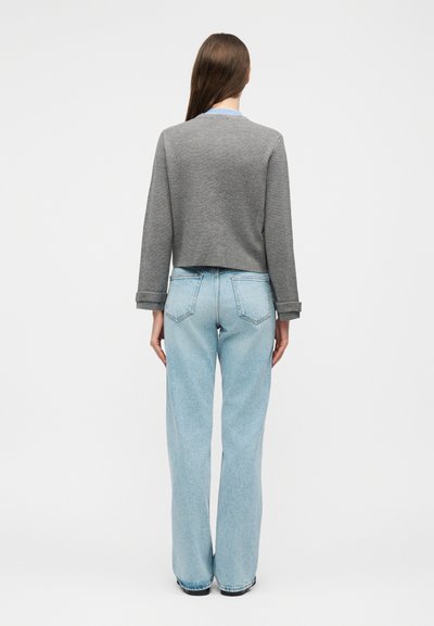 Γυναίκα που στέκεται με την πλάτη, φοράει γκρι μακρυμάνικη μπλούζα, ανοιχτό μπλε τζιν ψηλόμεσο με φαρδιά μπατζάκια και μαύρα παπούτσια σε λευκό φόντο.