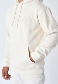 Sudadera color crema hecha de tela suave, con un bolsillo delantero, capucha con cordones y un diseño de corazón en relieve en el pecho.