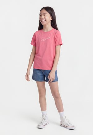 Fată tânără cu păr lung și întunecat, purtând un tricou roz Nike, pantaloni scurți din denim, șosete albe și teniși albi cu glezna înaltă, zâmbind și stând în picioare.