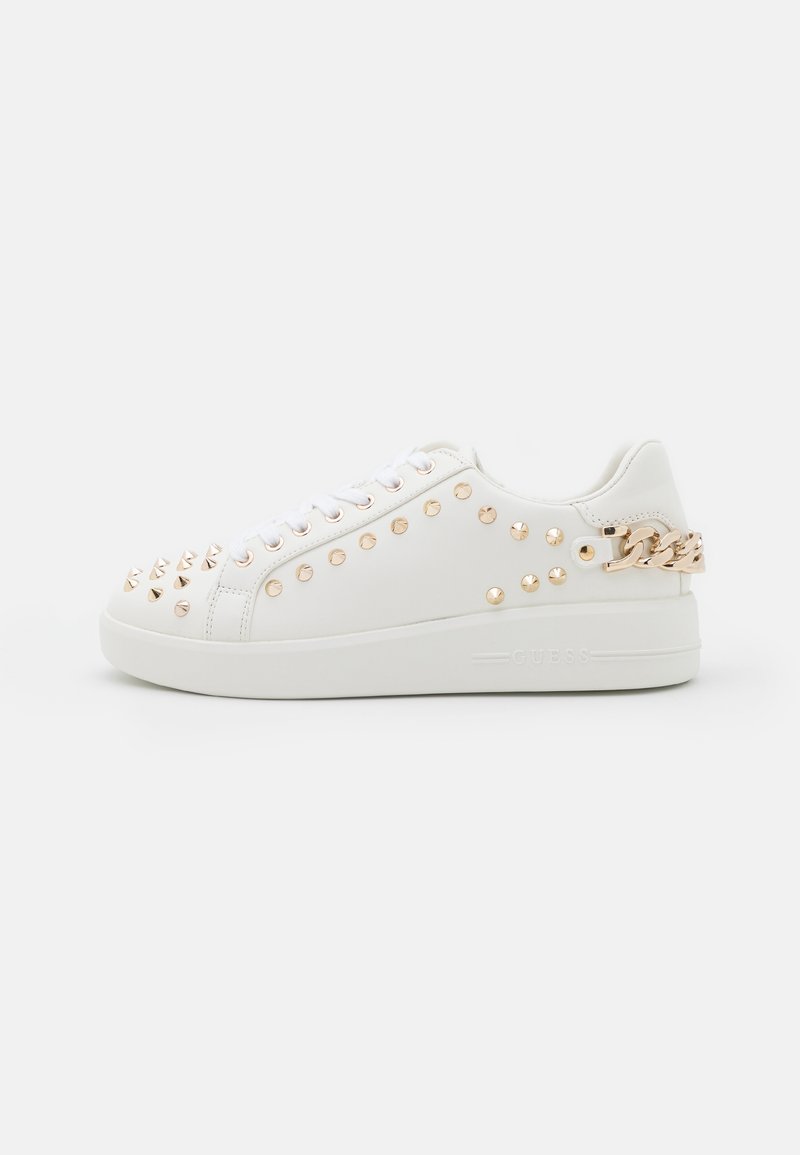 Witte lage sneaker met goudkleurige studs aan de voorkant en zijkanten, een gouden kettinkdetail op de hiel en een platte witte zool.