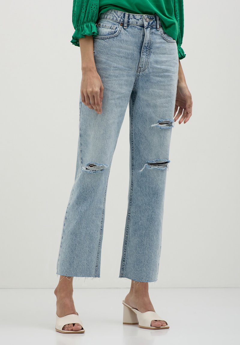 Femme portant un jean droit déchiré bleu clair, un blouson en œillets vert et des mules blanches à talons ouverts se tenant sur un sol blanc.