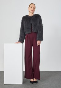 Mujer de pie con una mano apoyada en un pedestal rectangular blanco, vistiendo una chaqueta de piel sintética negra, pantalones de pierna ancha burdeos y tacones negros.