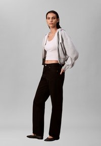 Grijze cropped hoodie, witte aansluitende tanktop, zwarte rechte broek en zwarte platte schoenen gedragen door een model tegen een grijze achtergrond.