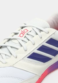 Balts sporta apavu pāris, izgatavots no tīkliņa un zamšāda; tam ir violetas svītras, pelēkas auklas un daudzkrāsains zolē ar rozā akcentiem.