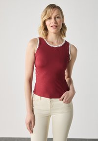 Rød ribbet tanktop med hvit skjortekant. Tett silhuett, uten ermer, kombinert med lysebeige bukser. Glatt tekstur.