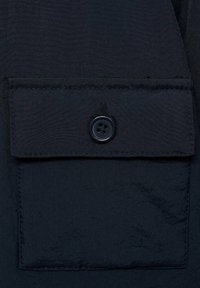 Navytyg nära håll som visar en fyrkantig ficka med en matchande knapp. Materialet verkar smidigt, med subtil textur och prydliga sömmar.