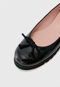 Ballerine en cuir verni noir avec un bout arrondi, ornée d'un nœud en corde noire torsadée et d'une doublure intérieure rose. Détails de semelle en caoutchouc visibles.
