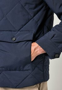 Marinblå kviltad jacka med en framficka med knappstängning, texturerad tyg och en strukturerad design. Lämplig för avslappnad klädsel.