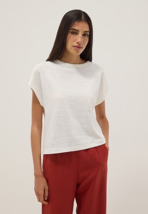 Jonge vrouw met lang donker haar, gekleed in een wit kort mouw shirt en een rode broek, staand met één hand in de zak tegen een effen achtergrond.