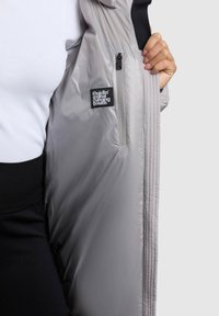 Lätt grå jacka med en ficka med dragkedja och synlig varumärkesetikett på det inre fodret, med en slät, glänsande yta.