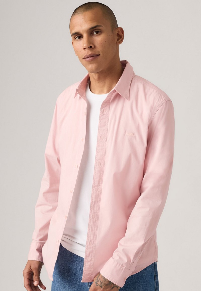 Camisa de botón rosa claro en una mezcla de algodón con cuello clásico y mangas largas, combinada con una camiseta blanca de cuello redondo.