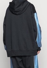 Svart hoodie med en blå satinsidpanel och snörning i huvan. Har ribbade muddar och nederkant samt en avslappnad passform.