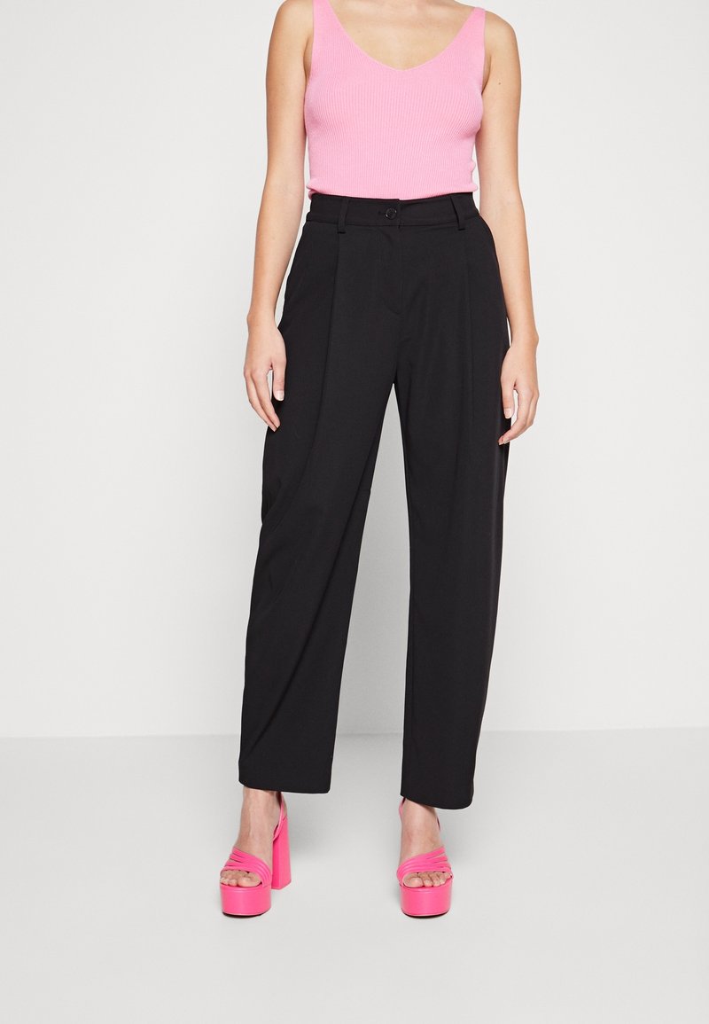 Pantalon noir large avec plis frontaux, associé à un débardeur côtelé rose et des talons compensés roses, mettant en valeur un contraste de couleurs vibrant.