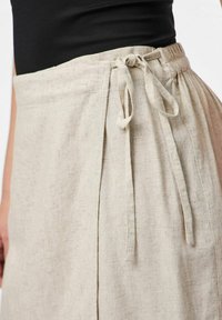 Beżowa lniana spódnica z wiązanym pasem, charakteryzująca się teksturowanym materiałem i bocznymi fałdami dla większej objętości. Prosty, funkcjonalny design o naturalnym wyglądzie.