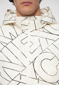 Homme portant un hoodie blanc avec des motifs linéaires abstraits noirs et des cordons de serrage, fermeture éclair fermée au niveau du cou, lèvres visibles mais yeux non montrés.