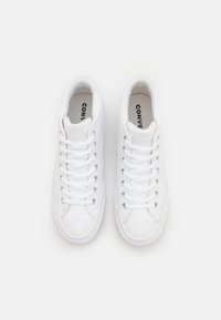 Zapatillas de lona blancas con un diseño de caña baja, que cuentan con cordones planos, ojales metálicos y una suela de goma texturizada. El logo de la marca es visible en la plantilla.