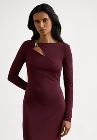 Burgundska obleka z dolgimi rokavi z zasukanim dizajnom in zlatim poudarkom na izrezu, izdelana iz gladkega, elastičnega materiala.