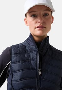 Marineblauw doorgestikt vest met een hoge kraag, ritssluiting en een subtiele glans. Gecombineerd met een aangesloten zwarte basislaag en een witte pet.