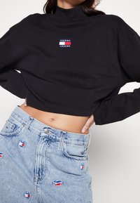 Black cropped turtleneck with "TOMMY JEANS" logo patch in red, blue, and white. Paired with high-waisted blue denim skirt featuring logo accents.