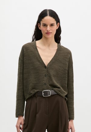 Femme aux longs cheveux foncés portant un cardigan vert olive boutonné à col en V et un pantalon marron taille haute avec une ceinture noire.