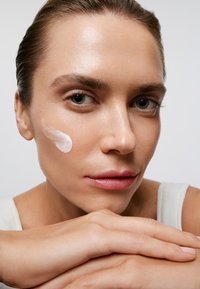 Hydratant sur la peau, texture lisse, couleur rose clair ; modèle avec des cheveux lisses, tenue neutre, reposant son menton sur ses mains contre un fond uni.