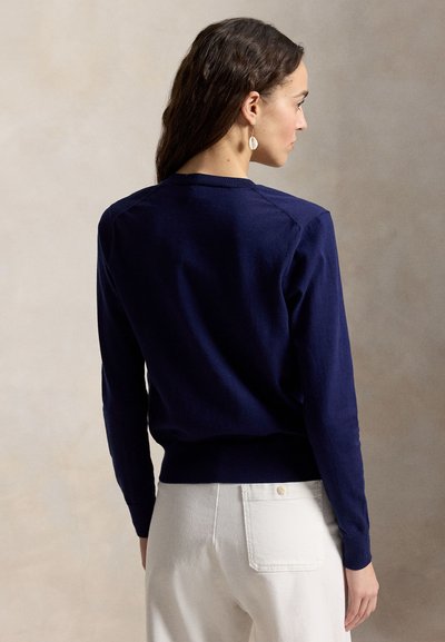 Sötétbarna hajú nő tengerészkék hosszú ujjú pulóvert és fehér nadrágot visel, hátulnézetből, semleges háttér előtt.