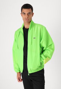 Casaco leve de cor verde néon, com gola alta, fecho de correr na frente e punhos elásticos. Apresenta um pequeno logotipo no lado esquerdo do peito e fecho com cordão.