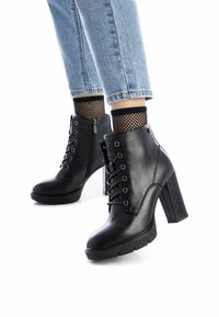 Botines negros con tacón alto; material de cuero, diseño de cordones, cremallera lateral y suela resistente. Llevados con jeans azules con vuelta y calcetines de red.