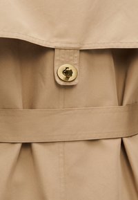Section de trench-coat beige avec une grande rabat, un bouton doré circulaire, une ceinture en tissu et un détail de jupe plissée. Texture lisse, design ajusté.