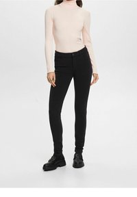 Femme portant un pull à col roulé rose clair, un pantalon noir ajusté et des mocassins noirs, debout devant un fond blanc uni.