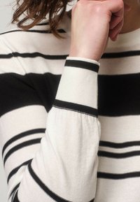 Pruhovaný svetr s tenkými černobílými vodorovnými proužky, žebrovanými manžetami, měkkou texturou a přiléhavým střihem na neutrálním pozadí.